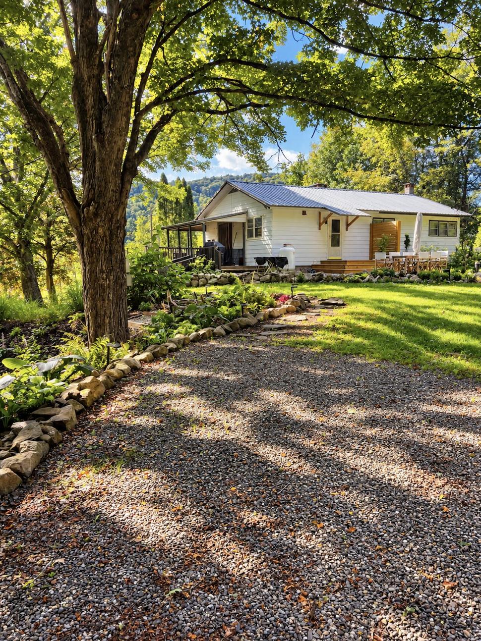 Ranch & Barn — the white clapboard ranch house under a standing-seam roof, seen down the gravel drive past stone-edged planting beds and a broad shade tree